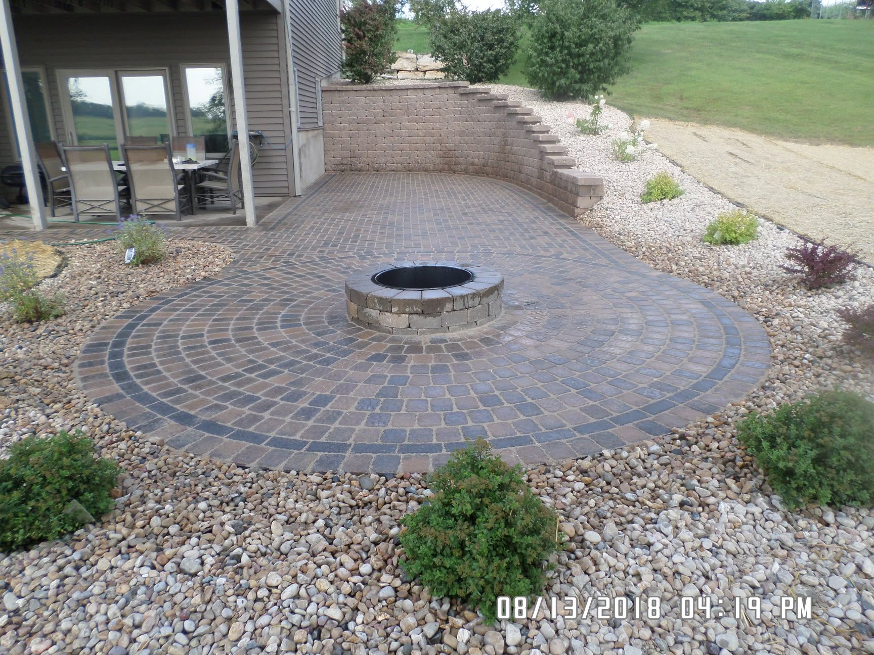 Circular paver patio with centered fire pit, seating wall, and river rock garden, Platteville WI