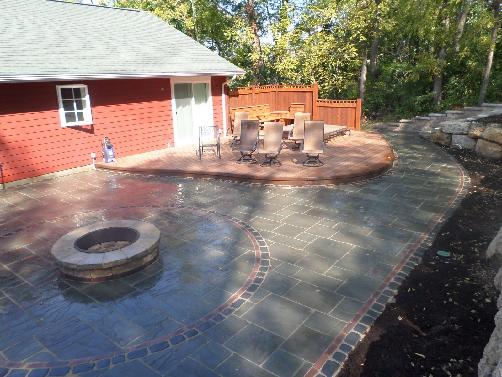 Stone paver patio with built-in fire pit, seating area, and wood deck overlooking a wooded backyard in Platteville, WI